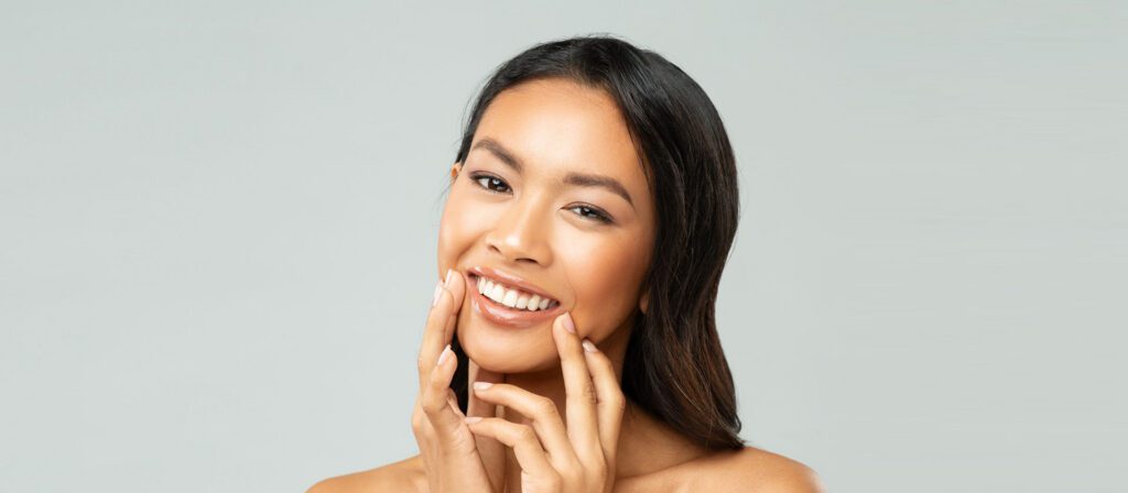 smiling-woman-with-dark-hair-resting-her-hand-near-her-face-against-a-gray-background